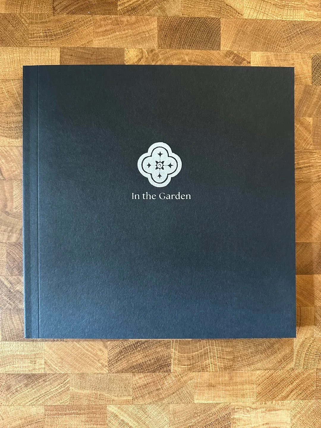 Black hardcover book titled In the Garden with a white floral emblem on a wooden surface