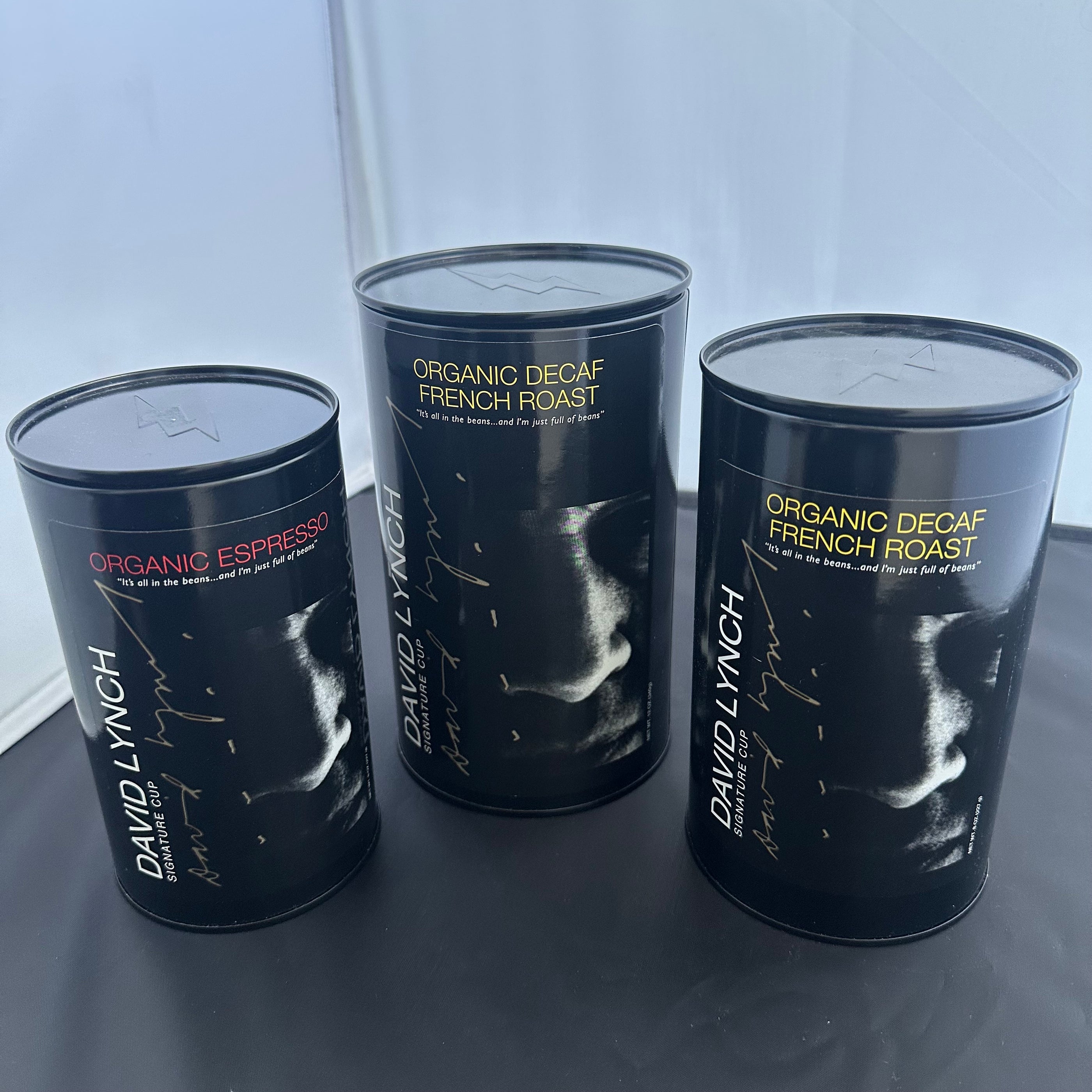 Three black coffee tins with artistic faces labeled David Lynch Coffee Tin, including Organic Espresso and Organic Decaf French Roast.